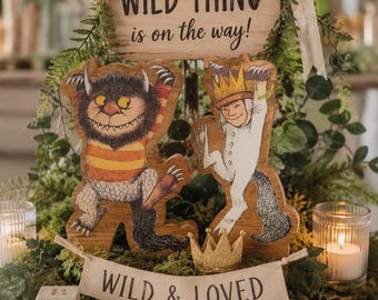 May include: A wooden sign reading a WILD THING is on the way! with two illustrated monster cutouts, a banner reading WILD & LOVED, a small gold crown, wooden alphabet blocks, and a pair of tan leather baby moccasins arranged on a wood slice.