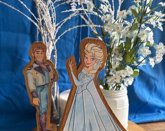 May include: Two wooden cutout figures featuring a male character in a light blue and grey uniform and a female character in a long light blue gown with a blonde braid. Both figures are mounted on oval wooden bases and stand upright.