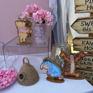 May include: A collection of party decorations featuring wooden cutouts of Winnie the Pooh, Eeyore, and Tigger, a jute rope beehive, a glass jar with pink flowers, a bowl of pink candies, and a wooden signpost with directional arrows.