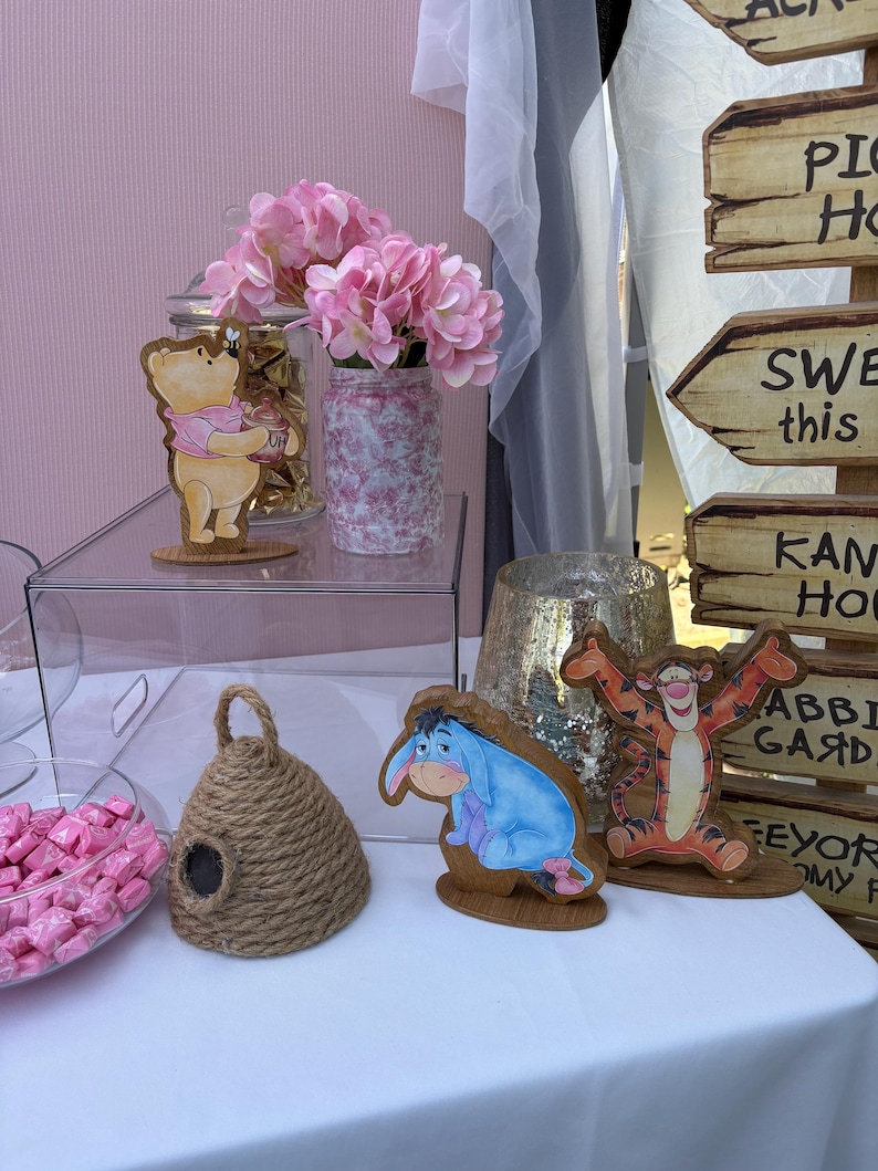 May include: A collection of party decorations featuring wooden cutouts of Winnie the Pooh, Eeyore, and Tigger, a jute rope beehive, a glass jar with pink flowers, a bowl of pink candies, and a wooden signpost with directional arrows.