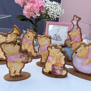 May include: A collection of six wooden Winnie the Pooh character cutouts featuring various poses, including one holding a honey pot labeled Hunny, one with a bee, and one resting on a pink balloon, all displayed on individual oval wooden bases.