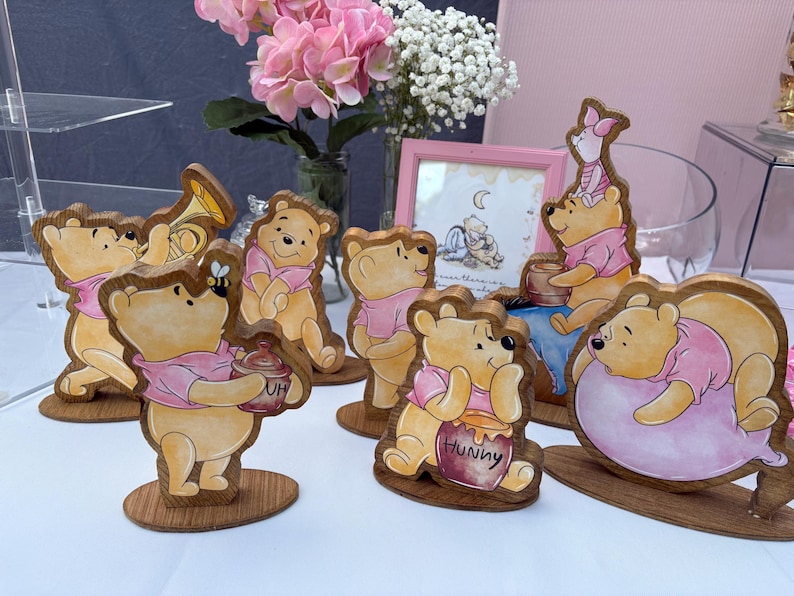May include: A collection of six wooden Winnie the Pooh character cutouts featuring various poses, including one holding a honey pot labeled Hunny, one with a bee, and one resting on a pink balloon, all displayed on individual oval wooden bases.