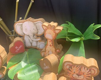 May include: Wooden animal figurines with heart-shaped signs that say "Be Strong", "Be Humble", and "Be Clever". The figurines include an elephant, giraffe, and tiger. The figurines are on a wooden base with a green leaf.