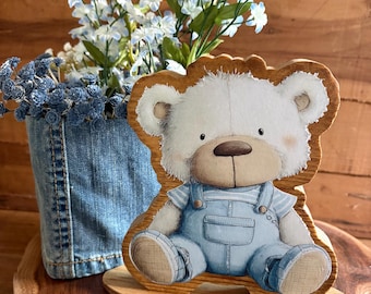May include: A wooden cutout of a white teddy bear wearing light blue denim overalls and a striped shirt. The bear is illustrated with a brown nose and sits on a flat oval wooden base. The cutout has a visible wood grain texture throughout.