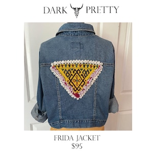 May include: A blue denim jacket with a triangular embroidered patch on the back. The patch features a yellow geometric design with red, green, and blue accents, surrounded by white seashells.