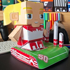 Könnte beinhalten: Eine Papierfigur einer Person in rot-weißer Uniform, die auf einer grünen Basis mit den Zahlen 26 und 50 steht. Die Figur hat blonde Haare und blaue Augen. Die Basis trägt den Text "BOX EDS PAPER CRAFTS".