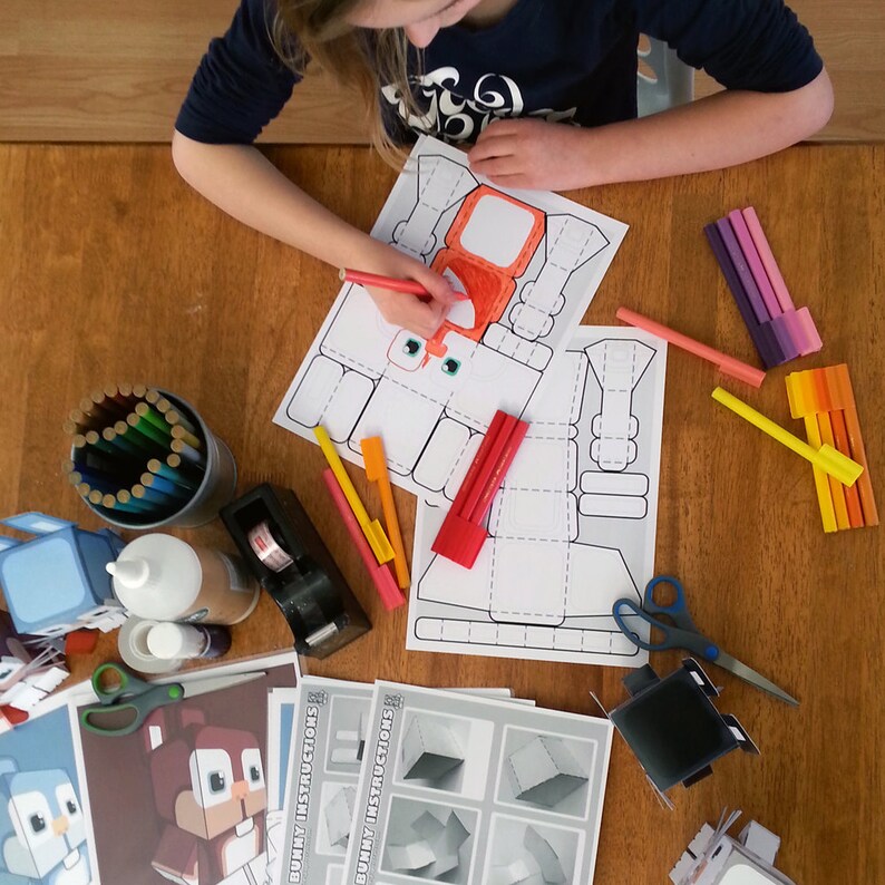 K&ouml;nnte beinhalten: Ein Kind malt ein Papiermodell eines Roboters mit roten, orangen und gelben Buntstiften aus. Der Roboter besteht aus Quadraten und Rechtecken. Das Papiermodell liegt auf einem Holztisch mit anderen Bastelutensilien, darunter Buntstifte, Kleber und eine Schere.
