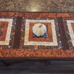 May include: A fall-themed table runner with a pumpkin centerpiece. The runner features three square quilted blocks with floral patterns in shades of brown, orange, and cream. The pumpkin is white and sits in the center block.