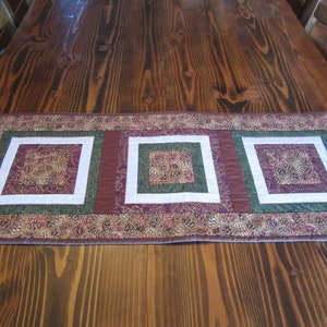 May include: A brown, green, and white quilted table runner with three square panels featuring a floral pattern. The runner is on a wooden table.