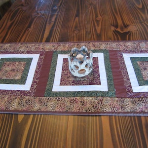 May include: A brown, green, and white quilted table runner with a decorative, white ceramic candle holder in the center.