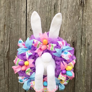 Puede incluir: Corona de Pascua con un conejito blanco de ganchillo, incluyendo orejas, patas y una cola esponjosa. La corona está adornada con malla morada, cintas coloridas y huevos de Pascua en colores pastel. Una decoración primaveral festiva.