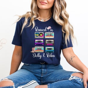May include: Navy blue t-shirt with a graphic design featuring cassette tapes and the text "Raised On Dolly & Reba." The design includes a cowboy hat and various cassette tape designs.