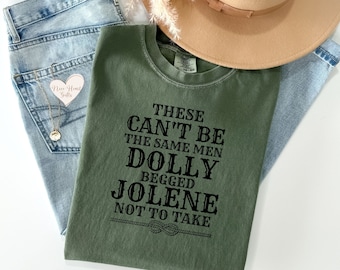 May include: An olive green t-shirt with the text "THESE CAN'T BE THE SAME MEN DOLLY BEGGED JOLENE NOT TO TAKE" in black. The shirt is paired with light wash denim jeans, a tan hat, and a gold necklace.