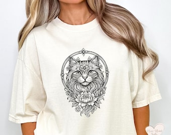 May include: Cream-colored t-shirt featuring a detailed black and white illustration of a cat's face within a decorative circular frame. The design includes floral elements and intricate linework, creating a whimsical and artistic aesthetic.