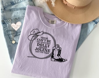 Pode incluir: Uma camiseta lavanda com um design gráfico preto que apresenta um chapéu de cowboy, um laço e uma bota de cowboy. O texto no gráfico diz "These can't be the same men Dolly begged Jolene not to take."