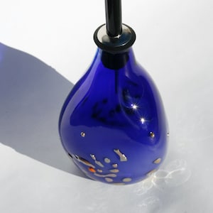 Könnte beinhalten: Eine kobaltblaue Glasflasche mit schwarzem Stopfen und silbernen Akzenten. Die Flasche hat eine perlmuttartige Oberfläche und goldene Flocken.