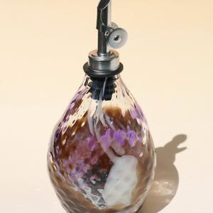 May include: A teardrop-shaped glass oil dispenser featuring a textured, dimpled surface with swirling patterns of brown, white, and purple. The bottle is topped with a silver-coloured metal pour spout and a black rubber seal at the neck.
