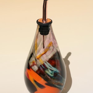 Peut inclure: Un distributeur d'huile en verre en forme de goutte d'eau avec un motif tourbillonnant de couleurs orange, bleu, blanc et brun foncé. La bouteille est surmontée d'un bec verseur en cuivre métallique et d'un bouchon en caoutchouc noir.