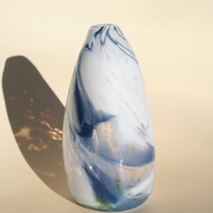 Peut inclure: Un vase en verre blanc et bleu avec un motif marbré tourbillonnant. Le vase a la forme d'une larme et une ouverture étroite.