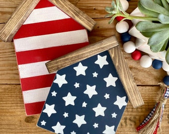 Op de afbeelding: Twee houten huisvormige borden versierd voor 4 juli. Een bord is rood-wit gestreept geverfd, het andere is blauw geverfd met witte sterren.