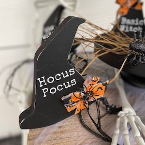 May include: Black wooden witch hat decoration with the words "Hocus Pocus" in white. The hat is adorned with an orange and black bow and a small spider. Halloween-themed decor.