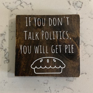 Peut inclure: Une pancarte en bois brun fonc&eacute; avec du texte blanc qui dit "Si vous ne parlez pas de politique, vous aurez de la tarte." Un contour blanc d'une tarte se trouve sous le texte.