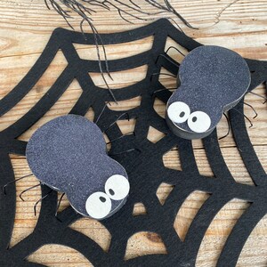 May include: Two black felt spiders with white eyes on a black felt spiderweb. The spiders are sitting on the web.