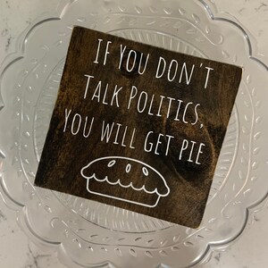 Peut inclure: Une pancarte en bois brun fonc&eacute; avec du texte blanc qui dit "Si vous ne parlez pas de politique, vous aurez de la tarte." Un contour blanc d'une tarte se trouve sous le texte.