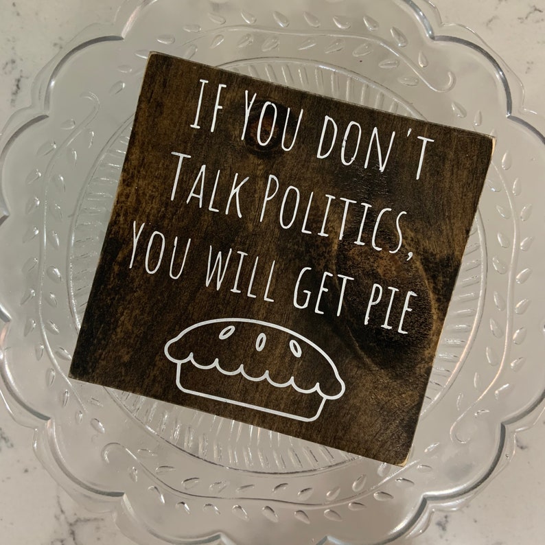 Peut inclure: Une pancarte en bois brun fonc&eacute; avec du texte blanc qui dit "Si vous ne parlez pas de politique, vous aurez de la tarte." Un contour blanc d'une tarte se trouve sous le texte.