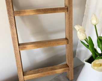 May include: A wooden ladder shelf with three shelves. The shelf is stained a dark brown color and has a rustic look. The shelf is sitting on a gray surface.