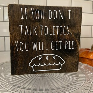 Peut inclure: Une pancarte en bois brun fonc&eacute; avec du texte blanc qui dit "Si vous ne parlez pas de politique, vous aurez de la tarte". Un contour blanc d'une tarte se trouve sous le texte.