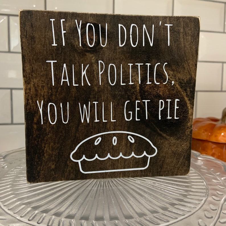 Peut inclure: Une pancarte en bois brun fonc&eacute; avec du texte blanc qui dit "Si vous ne parlez pas de politique, vous aurez de la tarte". Un contour blanc d'une tarte se trouve sous le texte.