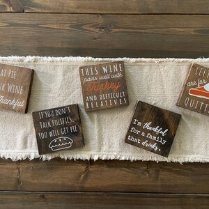 Peut inclure: Cinq pancartes en bois avec des dictons sur le th&egrave;me de Thanksgiving. Les pancartes sont peintes en brun avec des lettres blanches. Les dictons incluent "Mangez de la tarte Buvez du vin Soyez reconnaissant", "Si vous ne parlez pas de politique Vous aurez de la tarte", "Ce vin se marie bien avec la dinde Et les parents difficiles", "Je suis reconnaissant pour une famille qui boit", et "Les restes sont pour les l&acirc;ches".
