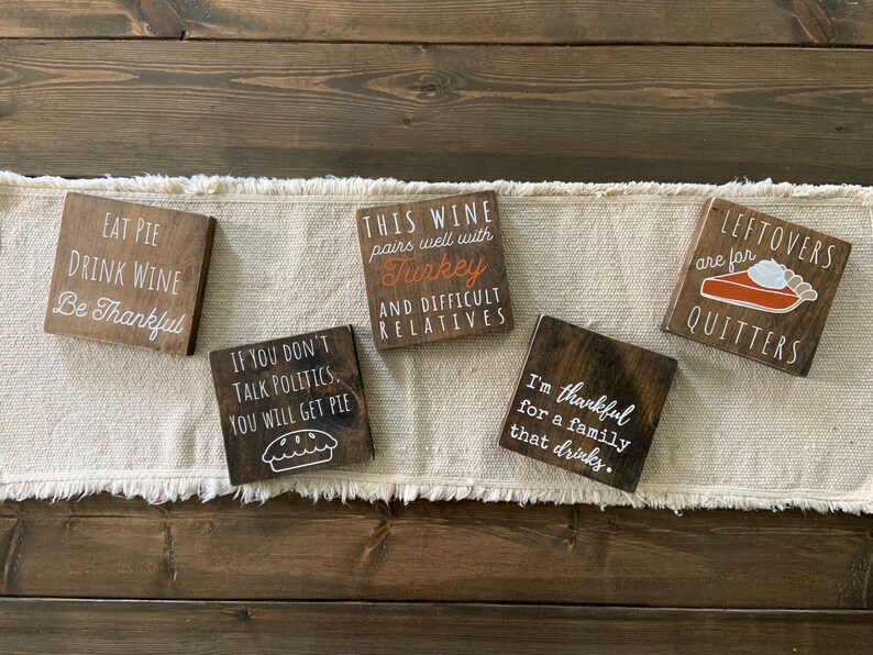 Peut inclure: Cinq pancartes en bois avec des dictons sur le th&egrave;me de Thanksgiving. Les pancartes sont peintes en brun avec des lettres blanches. Les dictons incluent "Mangez de la tarte Buvez du vin Soyez reconnaissant", "Si vous ne parlez pas de politique Vous aurez de la tarte", "Ce vin se marie bien avec la dinde Et les parents difficiles", "Je suis reconnaissant pour une famille qui boit", et "Les restes sont pour les l&acirc;ches".