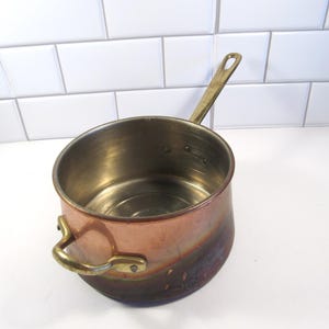 May include: A copper saucepan with a brass handle and a smaller brass handle on the side. The interior of the pan is a shiny silver color. The exterior of the pan has a gradient of colors, from copper to a darker brown.