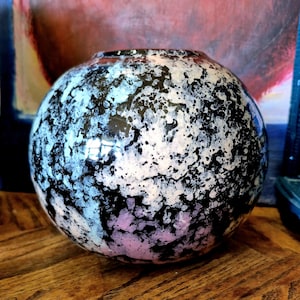 Puede incluir: Un jarrón de cerámica redondo con un patrón moteado en negro, blanco y azul claro. El jarrón tiene una abertura ancha y está sobre una superficie de madera. El diseño es abstracto y artístico.