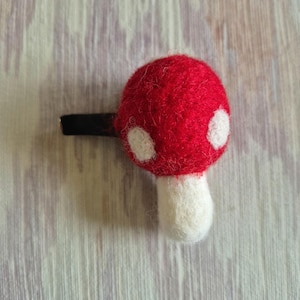 May include: A red mushroom hair clip with white spots and a white stem. The clip is made of felt and has a black barrette. This whimsical accessory is perfect for adding a touch of nature-inspired charm to any hairstyle.