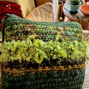Puede incluir: Un cojín rectangular hecho a mano con un diseño texturizado y multicolor. El cojín presenta bandas de hilo verde oscuro, verde oliva y verde amarillento, con una franja rizada texturizada en el centro. Un ribete decorativo de color dorado acentúa la parte inferior.