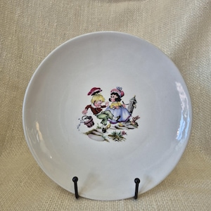 May include: White ceramic plate featuring a colorful illustration of two children playing. The children are dressed in vintage clothing and are surrounded by flowers and a small basket. The plate is displayed on a black metal stand.