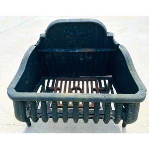 May include: A dark green cast iron fireplace grate featuring a high back panel with a moulded fleur-de-lis emblem. The structure includes vertical front bars and a central rectangular metal grate insert with visible rust and weathered texture.