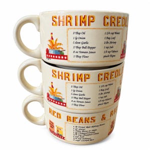 May include: Three stacked cream-colored ceramic mugs featuring printed recipes. The top two mugs display a Shrimp Creole recipe with ingredients and instructions. The bottom mug features a Red Beans and Rice recipe with text and illustrations.