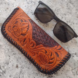Puede incluir: Un estuche para gafas de cuero marrón con un diseño floral y costuras negras. El estuche está sobre una superficie gris junto a unas gafas de sol negras.
