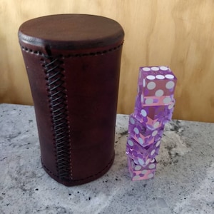 Può includere: Un bicchiere da dado in pelle marrone con una cucitura e una pila di cinque dadi viola con punti bianchi.