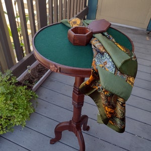 May include: A vintage wooden side table with a round, green felt-covered top. A decorative scarf with paisley and floral patterns in shades of green, orange, and gold is draped over the table. A small, hexagonal wooden box sits on the table.