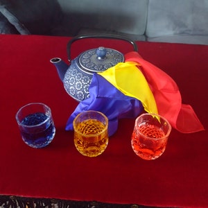 Könnte beinhalten: Eine blaue Teekanne mit Blumenmuster steht auf einem roten Tisch mit drei Gläsern, die mit blauen, gelben und roten Flüssigkeiten gefüllt sind. Ein farbenfroher Seidenschal ist über die Teekanne gelegt.