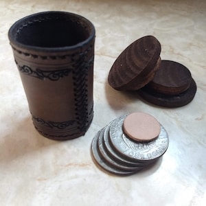 Op de afbeelding: Een bruine leren beker met decoratieve stiksels en een stapel zilveren munten met een klein, rond, bruin lederen stukje erop. Ook zijn er drie houten schijven.