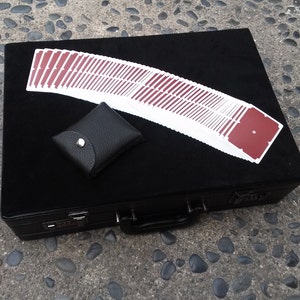 May include: A black case with a handle and a combination lock, a deck of playing cards with red backs, and a small black leather coin purse with a snap closure. The cards are fanned out on top of the case.