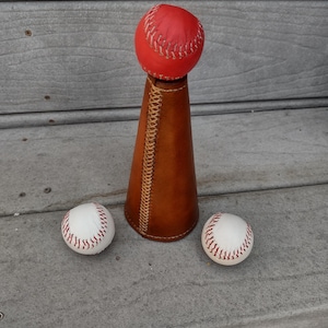 May include: A baseball-themed decorative item featuring a red baseball atop a brown leather cone, with two additional baseballs. The cone is stitched with a lighter thread. The items are displayed on a gray wooden surface.