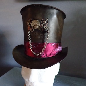 May include: A black leather top hat with a pink and floral patterned band. The hat features a silver chain and a steampunk-style gear design on the front.