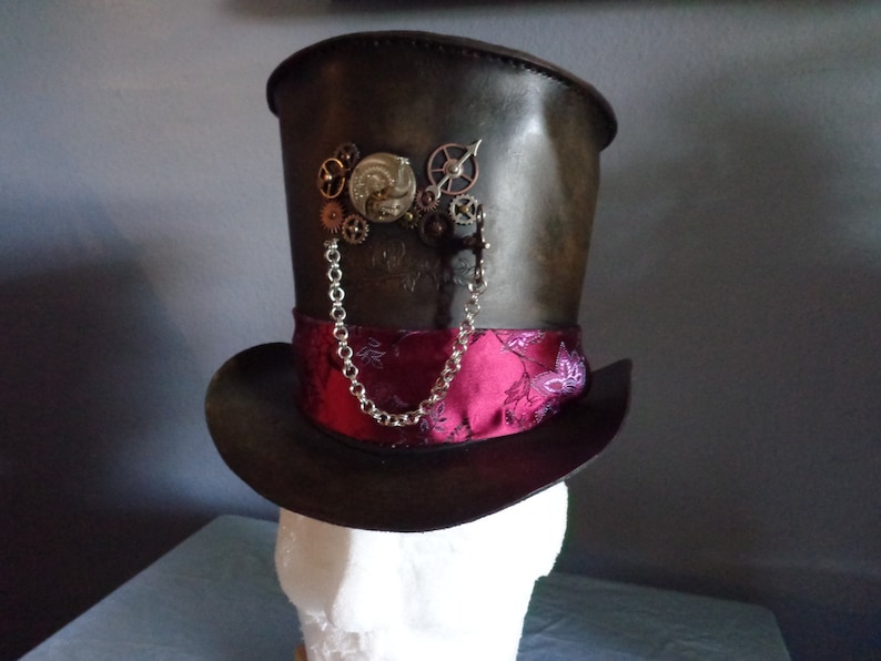 May include: A black leather top hat with a pink and floral patterned band. The hat features a silver chain and a steampunk-style gear design on the front.